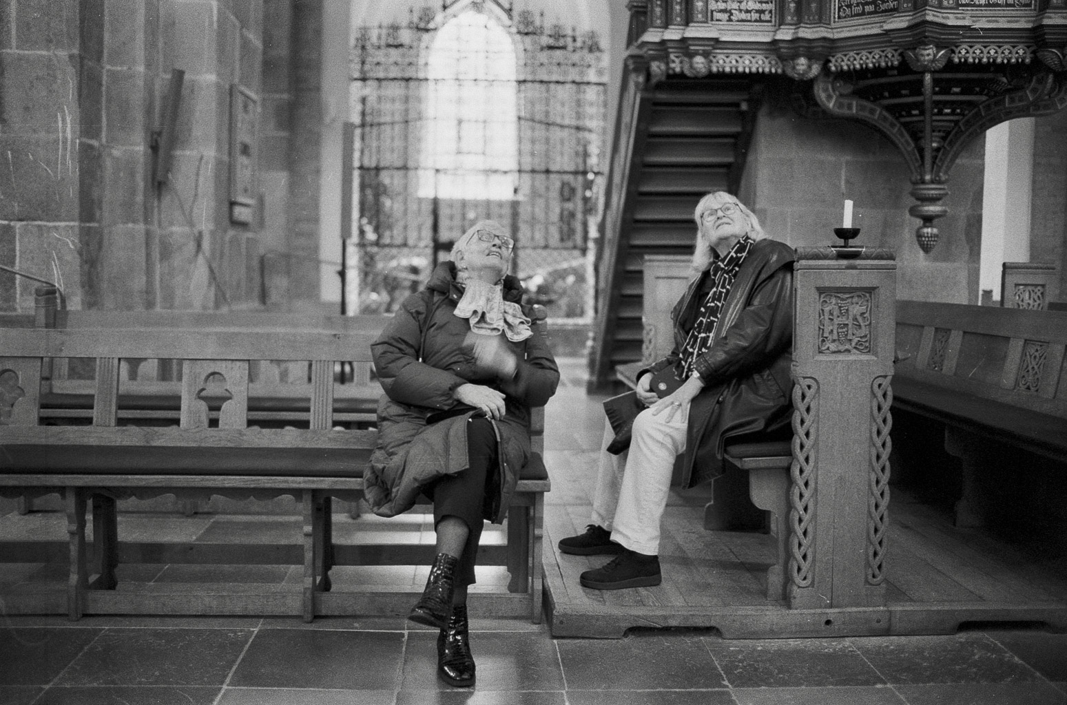 Two elderly individuals sitting inside a church, looking up with contemplative expressions. The interior features wooden benches and ornate architecture, with a large window letting in light.