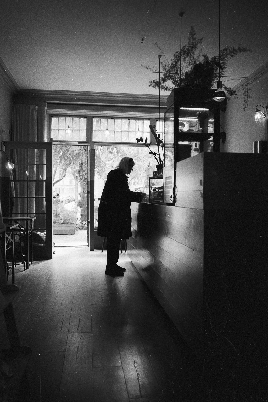A black and white image of a person standing at a counter in a café, with large windows and plants visible in the background.