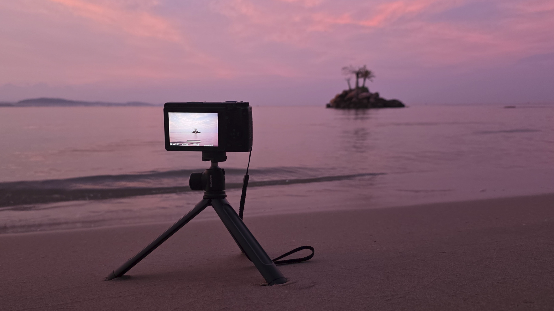 A camera on a tripod on a sandy beach, capturing a serene sunset over a calm sea with a small island in the background.