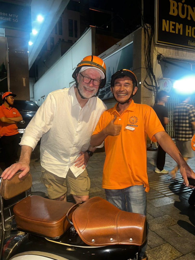 A smiling man with glasses and a beard poses next to a cheerful man in an orange shirt and helmet, in front of a vintage motorcycle at night in an urban setting.