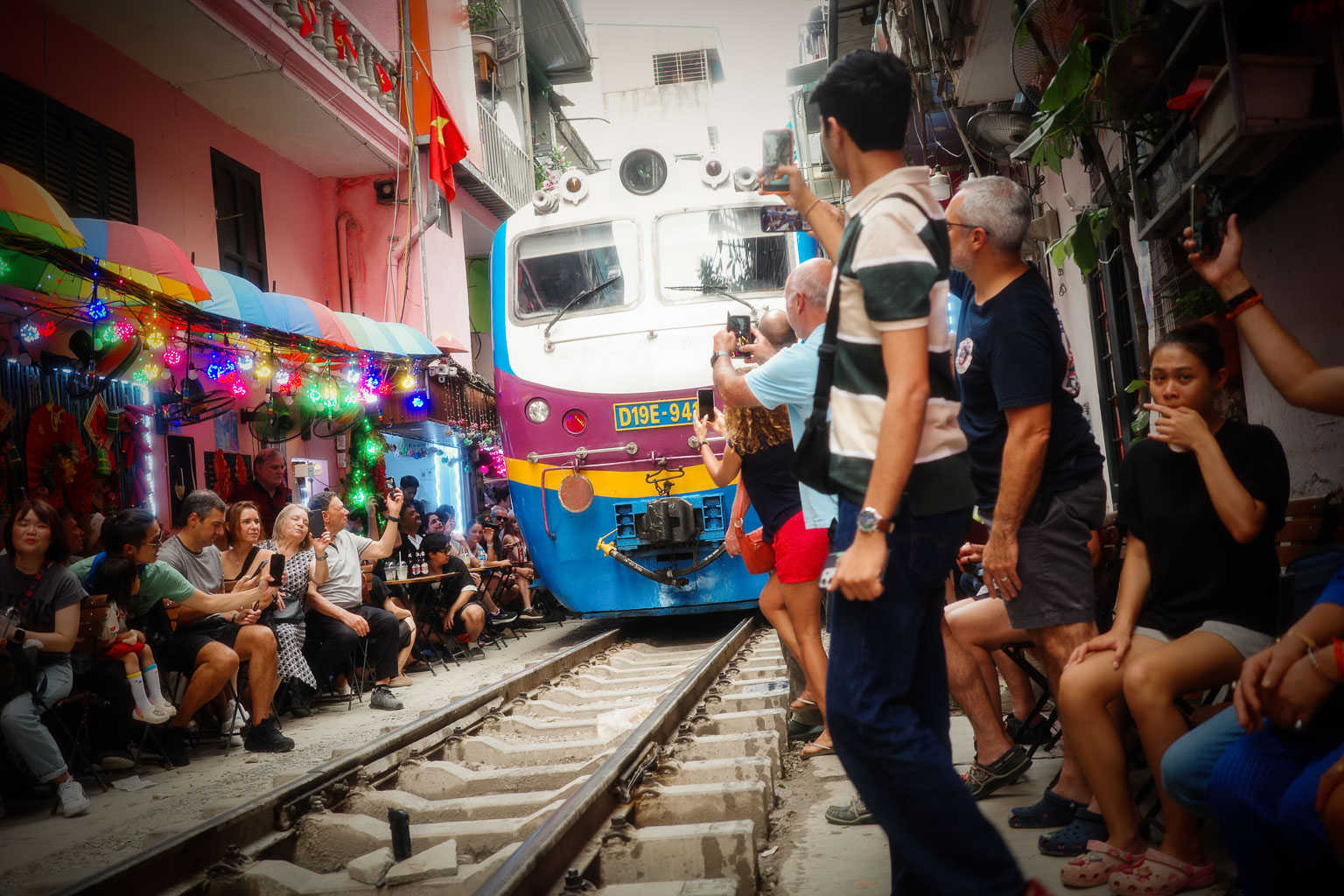 A colorful train approaching a busy market street, with people seated and taking photos beside the railway tracks, surrounded by vibrant decorations and lights.