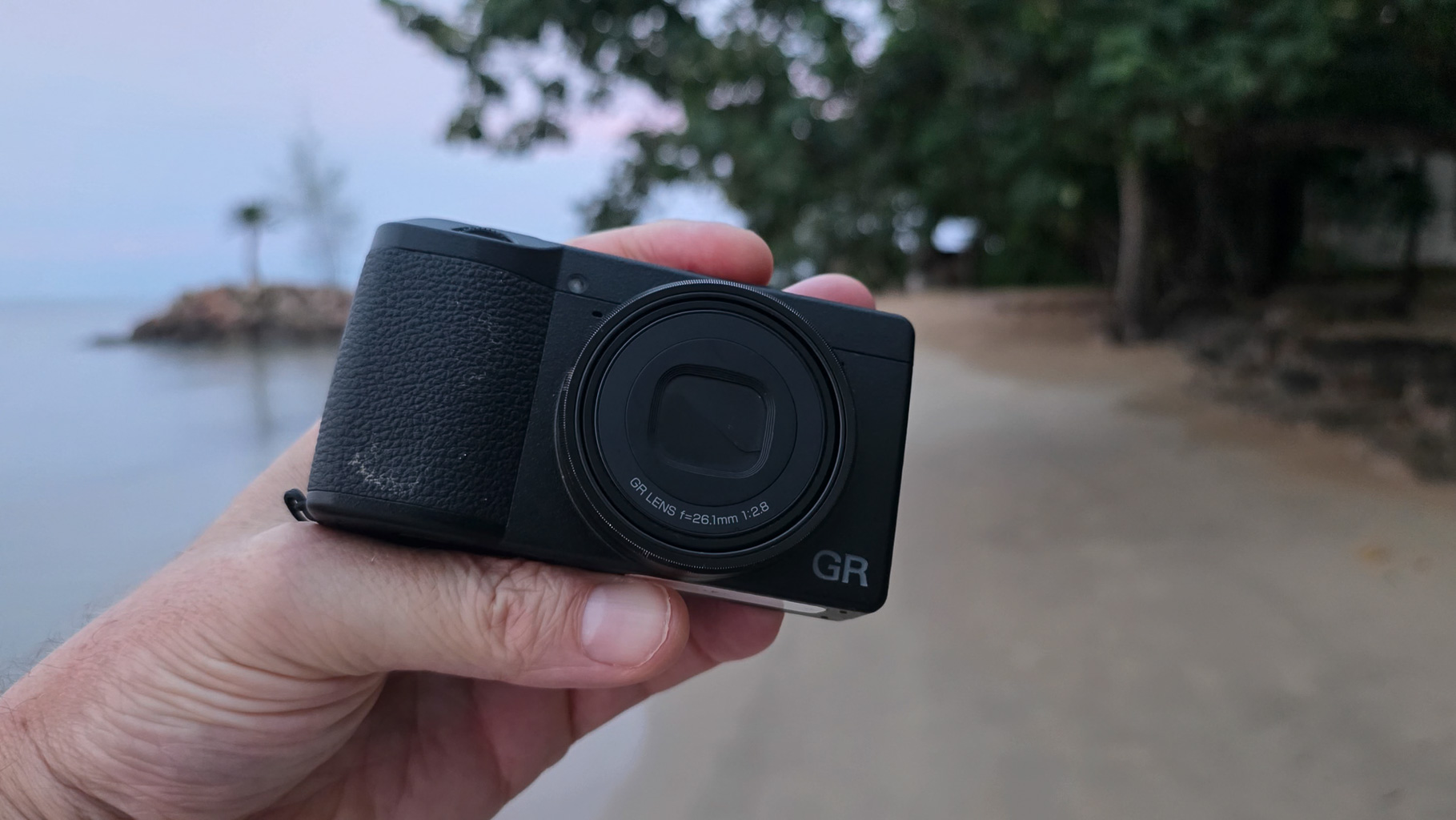 A person holding a compact digital camera with a black body, positioned against a blurred beach background during dusk.