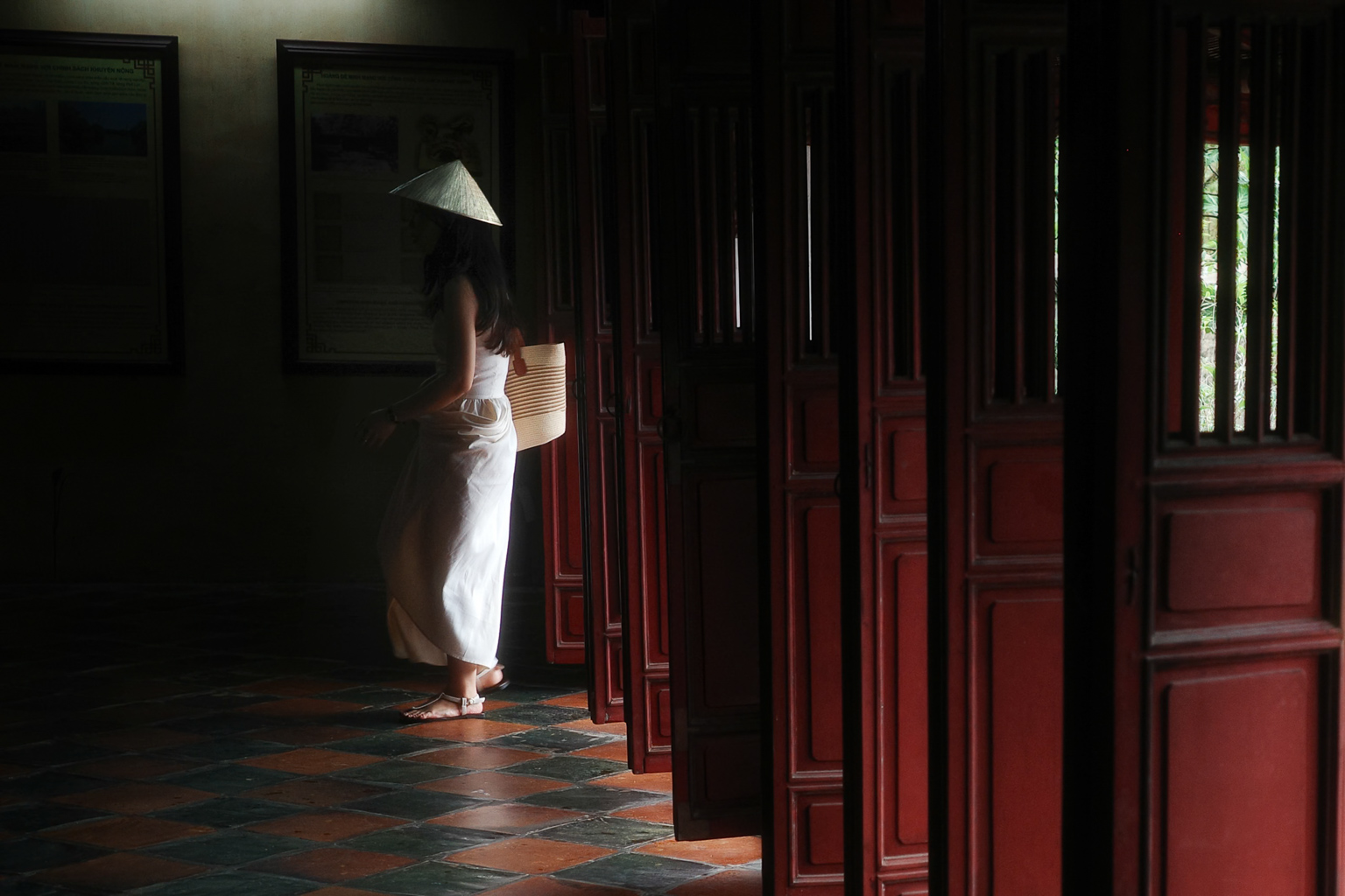 A woman wearing a traditional conical hat stands in a dimly lit corridor with red wooden doors, holding a woven basket.