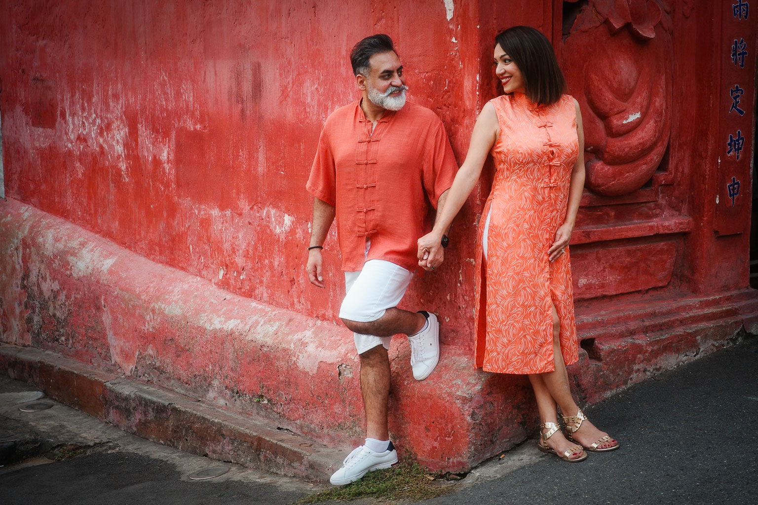A couple dressed in matching orange outfits standing hand in hand against a vibrant red wall, smiling at each other.