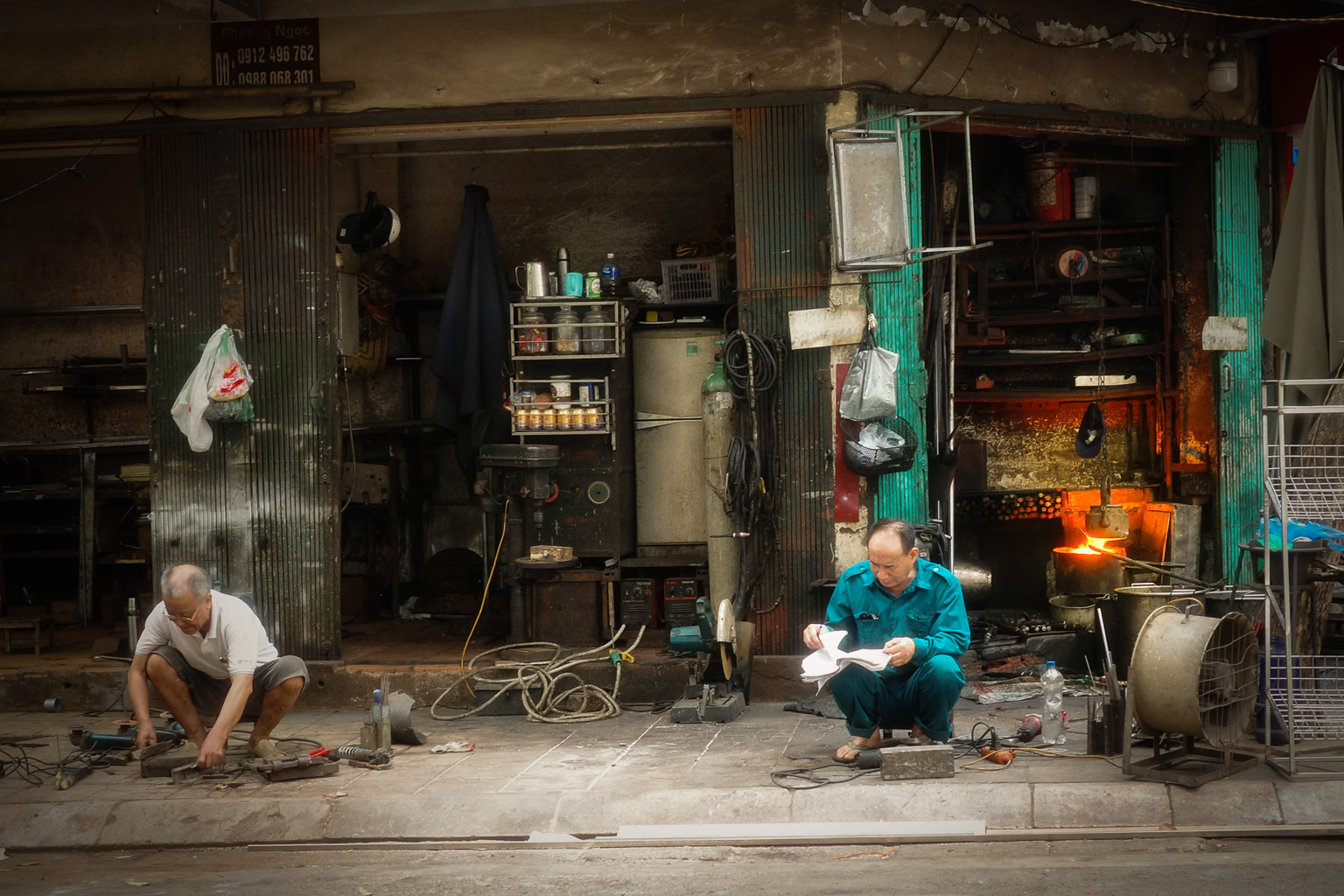Two men working in front of a workshop, one repairing items on the ground and the other reading a document while sitting.