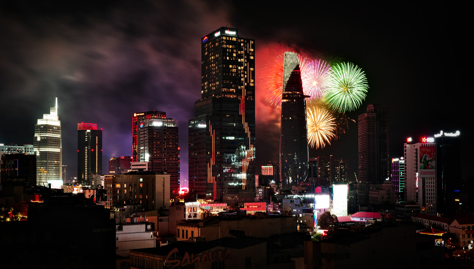 City skyline at night featuring tall buildings with vibrant lights and fireworks in the background.