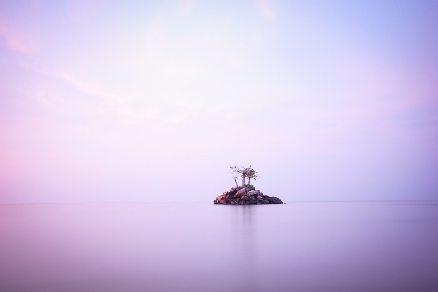 A small rocky island with palm trees surrounded by calm water, under a pastel-colored sky at dusk.