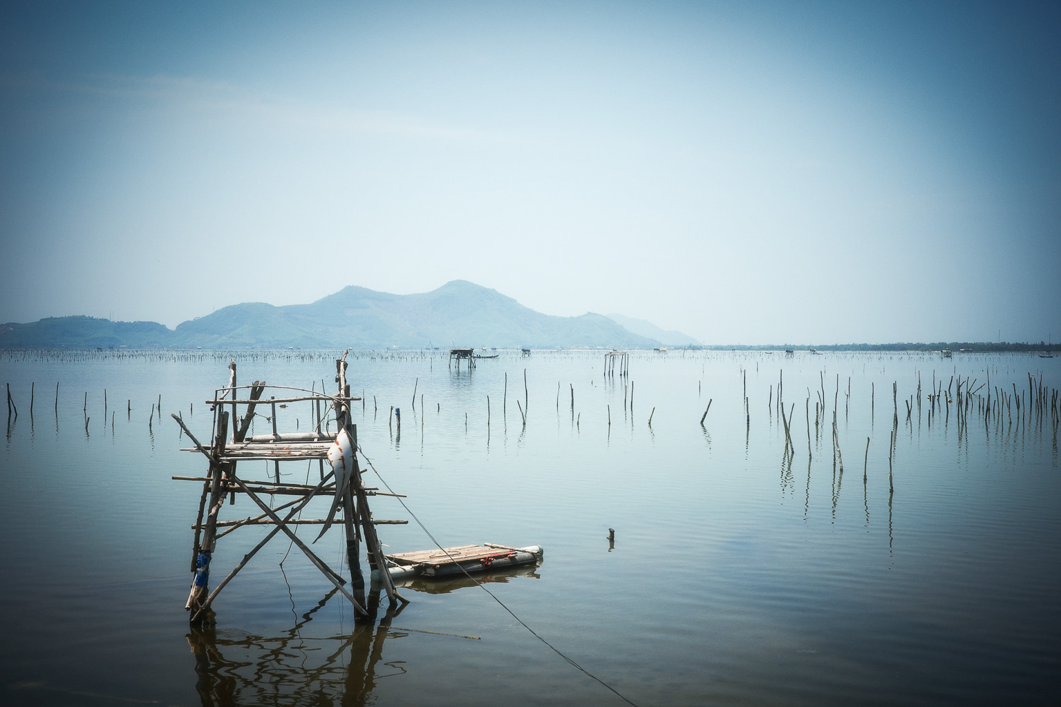 A wooden structure stands by a serene lake, with distant mountains in the background and vertical stakes protruding from the water.
