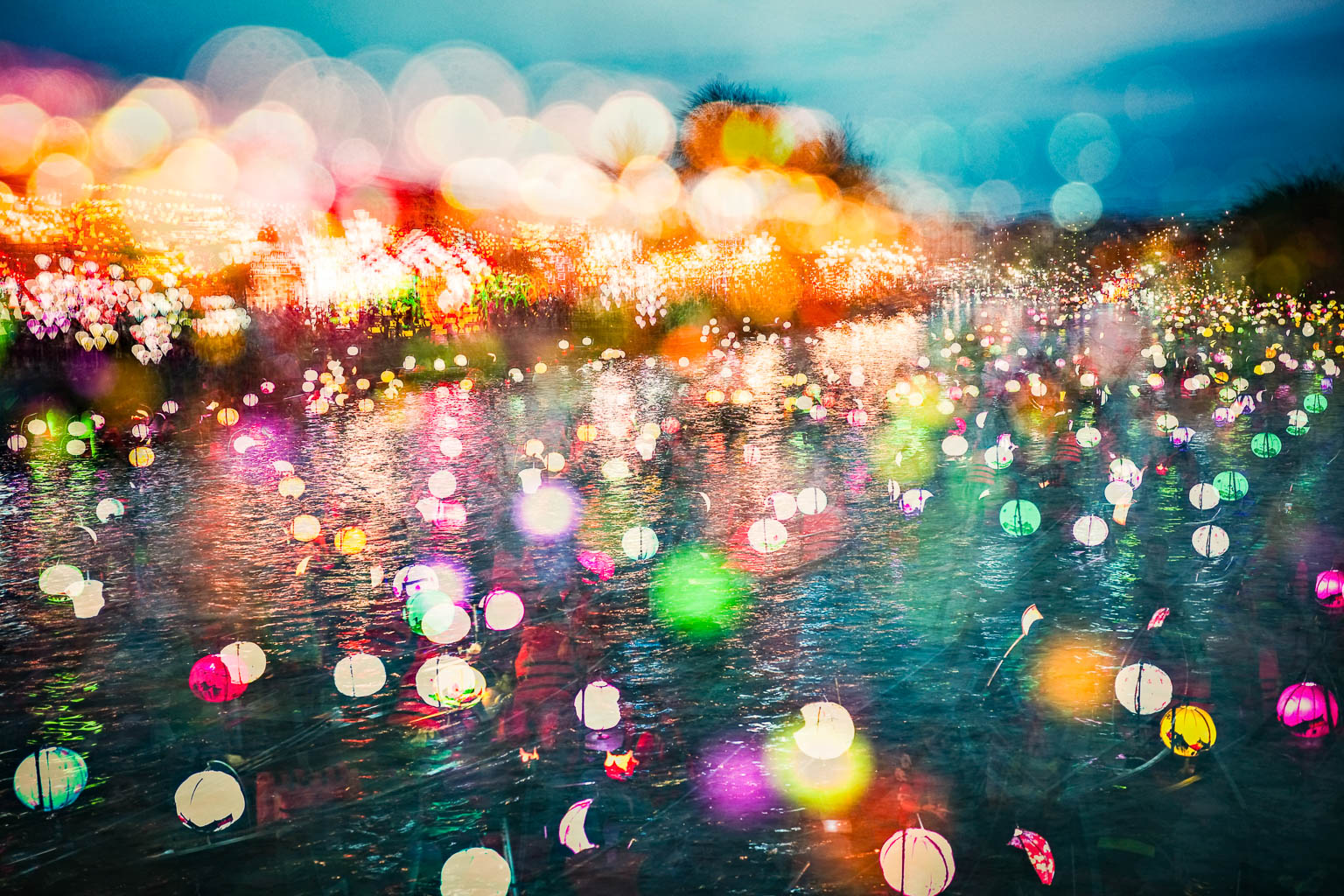 A colorful display of illuminated lanterns floating on a body of water, surrounded by a blurred, twinkling background of lights.