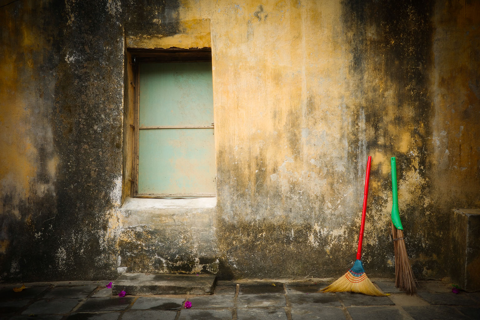 A weathered wall with a partially open window and two brooms, one red and one green, leaning against the wall. The floor is made of stone tiles, and there are a few scattered flower petals.