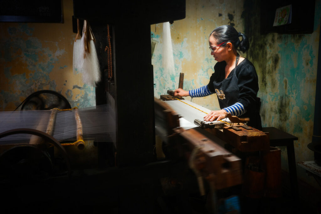 A woman weaving fabric on a traditional loom in a dimly lit workshop with textured, peeling walls.