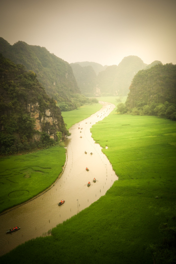 A scenic view of a winding river surrounded by lush green fields and towering limestone mountains, with small boats visible on the water under a misty sky.