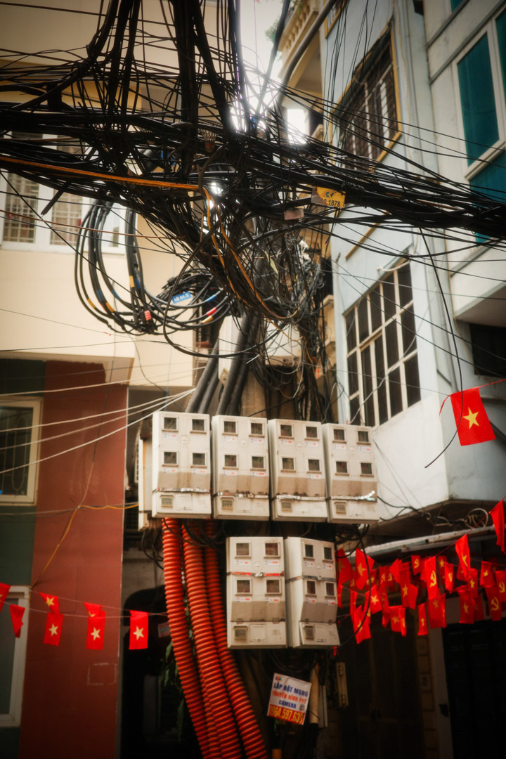 Chaotic arrangement of electrical wires above several utility meters in an urban alleyway, decorated with small red flags.