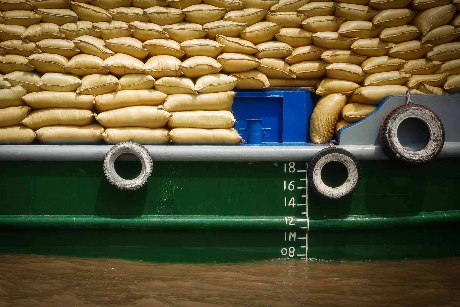 Close-up of a green boat loaded with stacked yellow sacks, featuring a measurement scale and two mooring tires.