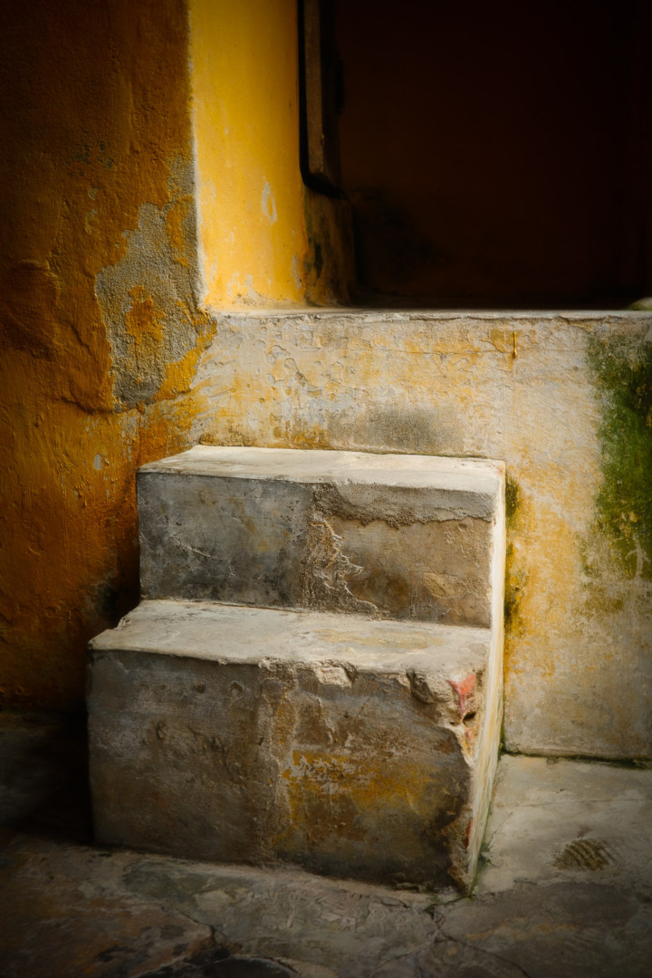 A weathered stone step with a textured surface, positioned against a yellow wall, leading up to a darkened doorway.