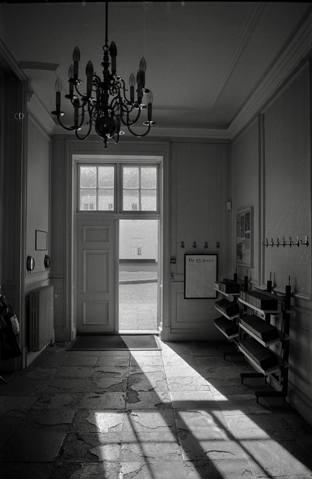 An interior view of an entrance hallway featuring a chandelier, a large door leading outside, and a wooden shelf with hooks and a notice board.