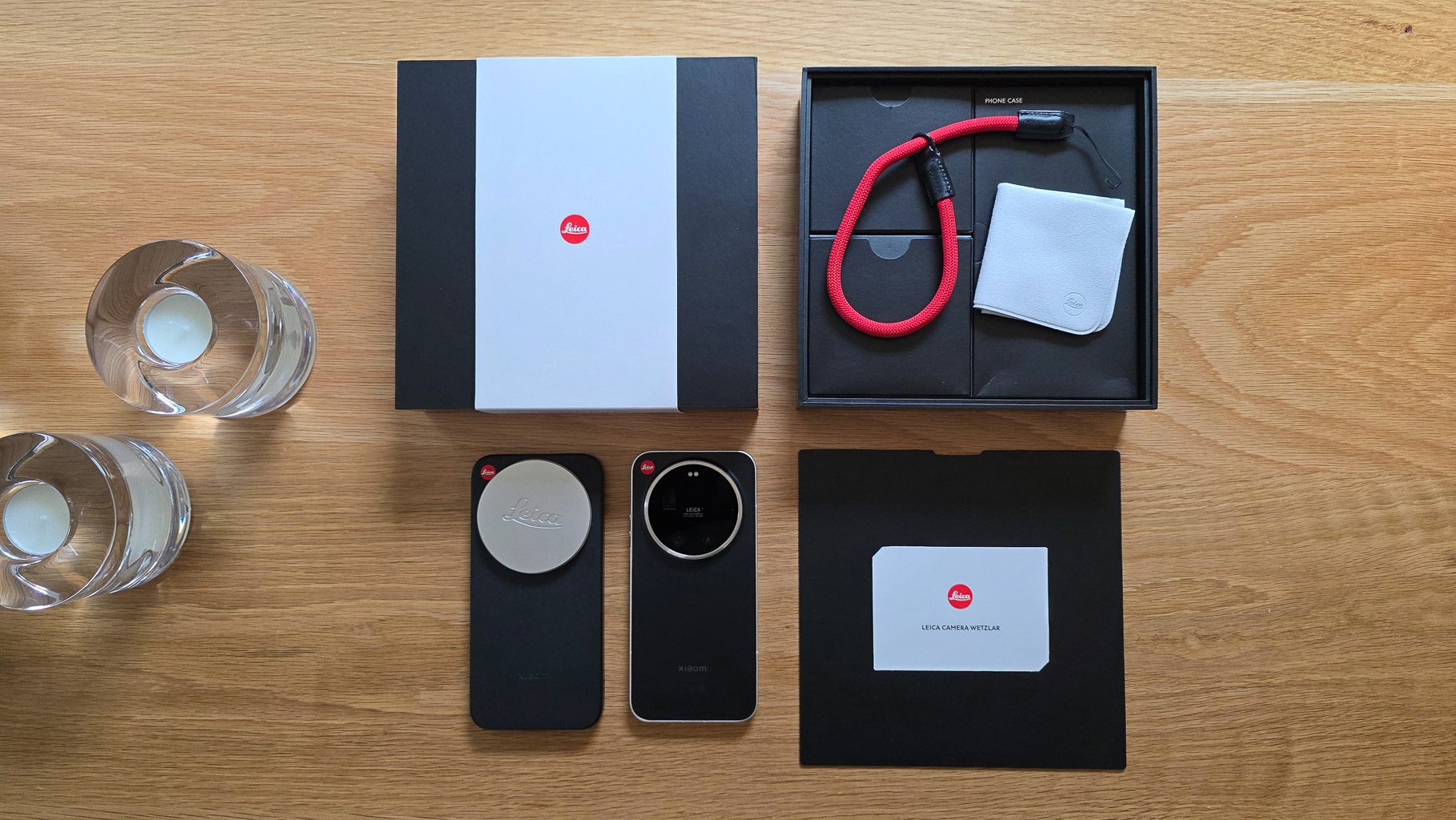 A flat lay of a Leica camera packaging. The image shows two camera lenses, a camera strap in red, a lens cap, a small white cloth, and a card, all placed on a wooden surface alongside two clear glasses with candles.