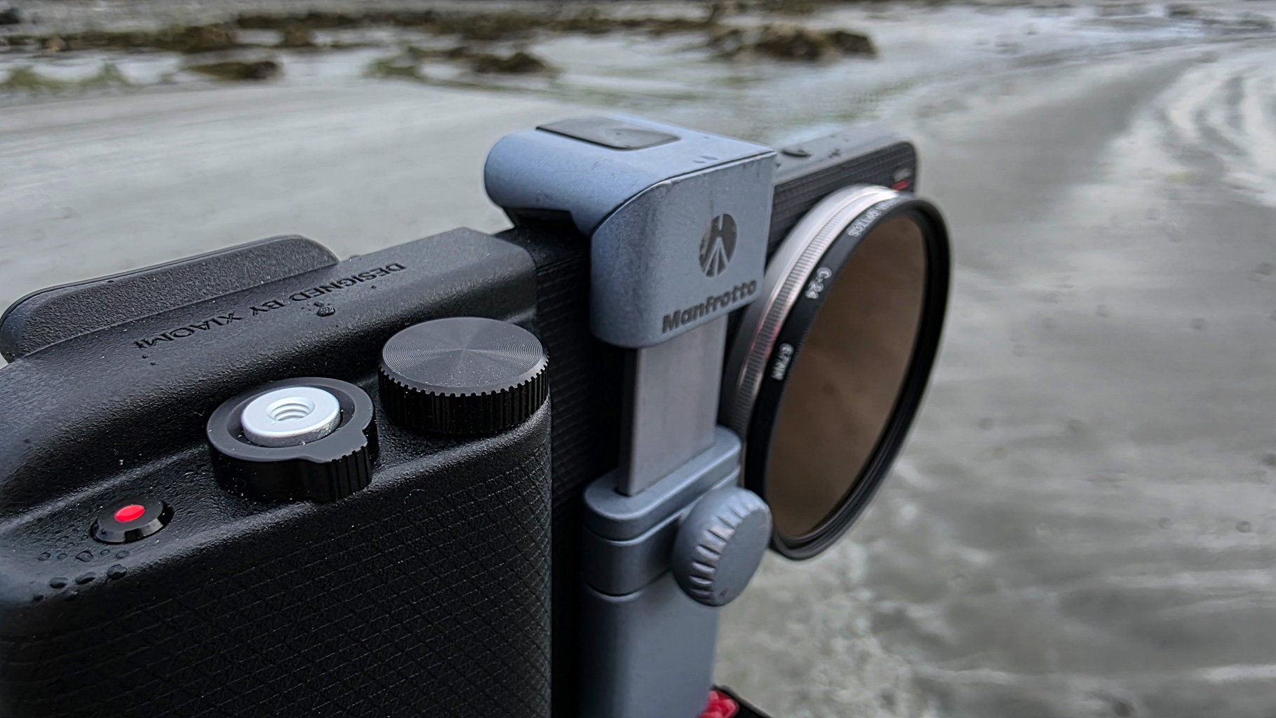 Close-up of a camera featuring a Manfrotto accessory, with visible controls and a neutral density filter, set against a blurred beach background.