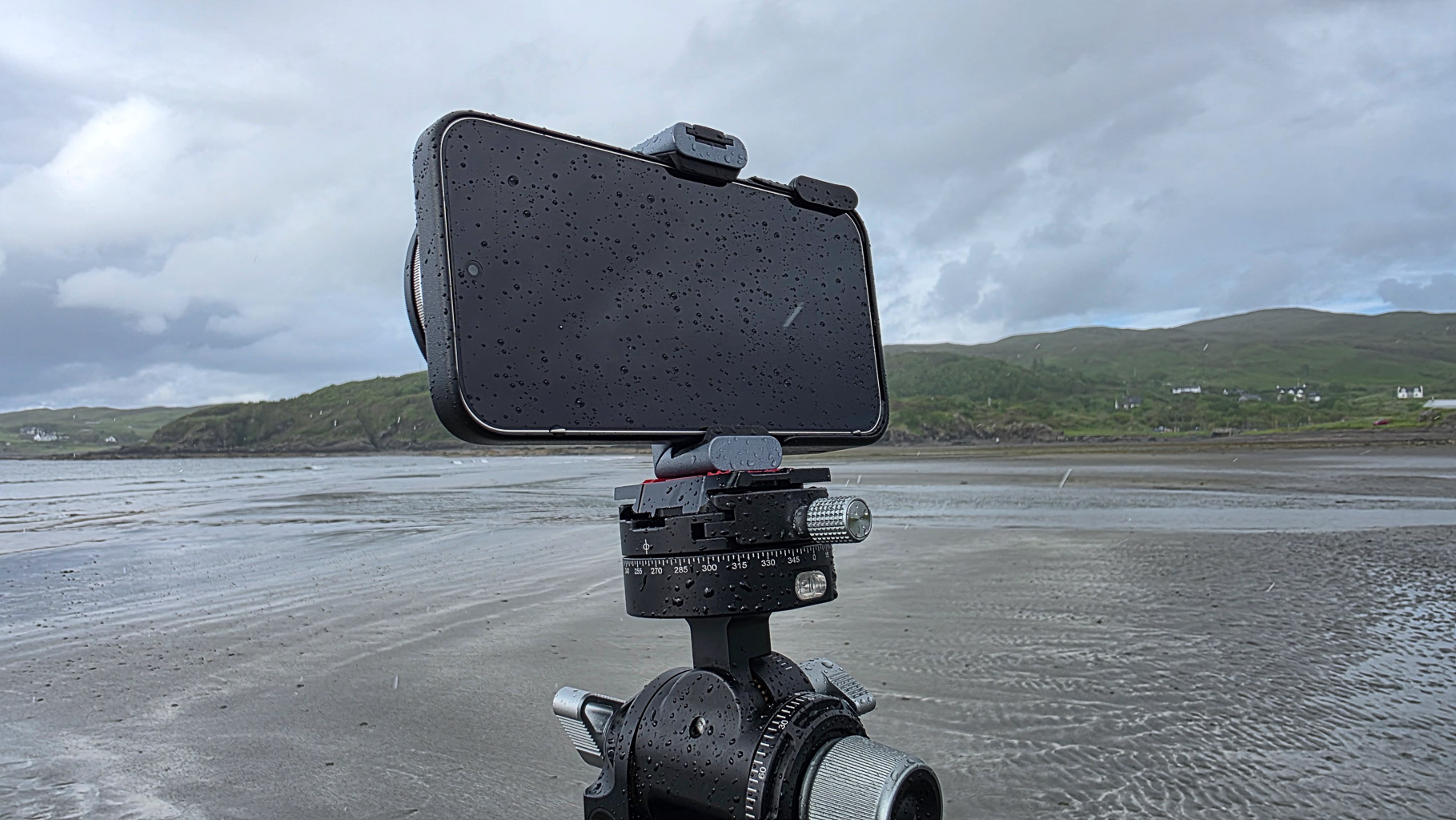 A smartphone mounted on a tripod with wet rain droplets on the screen, set against a cloudy beach landscape with rolling hills in the background.