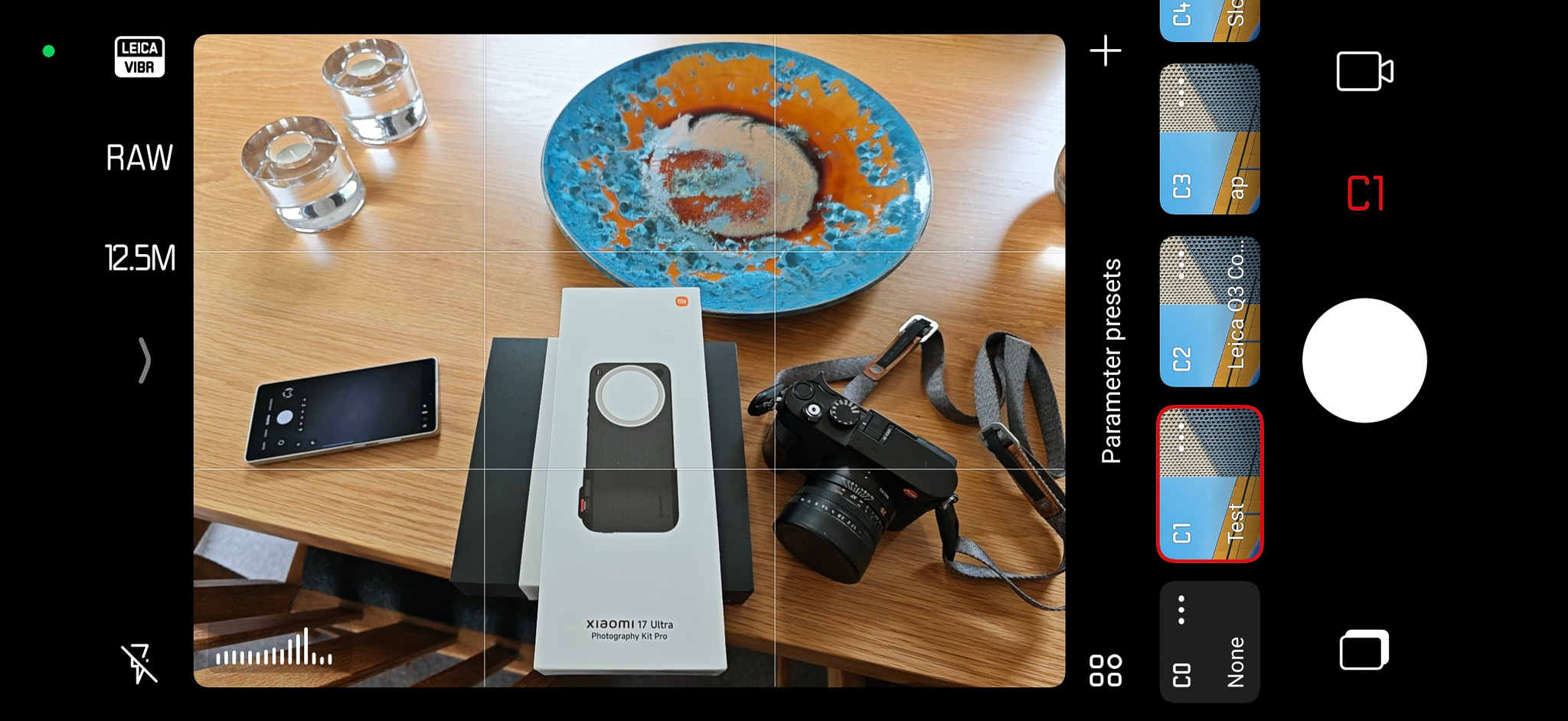 A tabletop scene featuring a Xiaomi 17 Ultra smartphone in a photography kit, a camera, a decorative blue and orange plate, and glass items, captured from a camera interface.