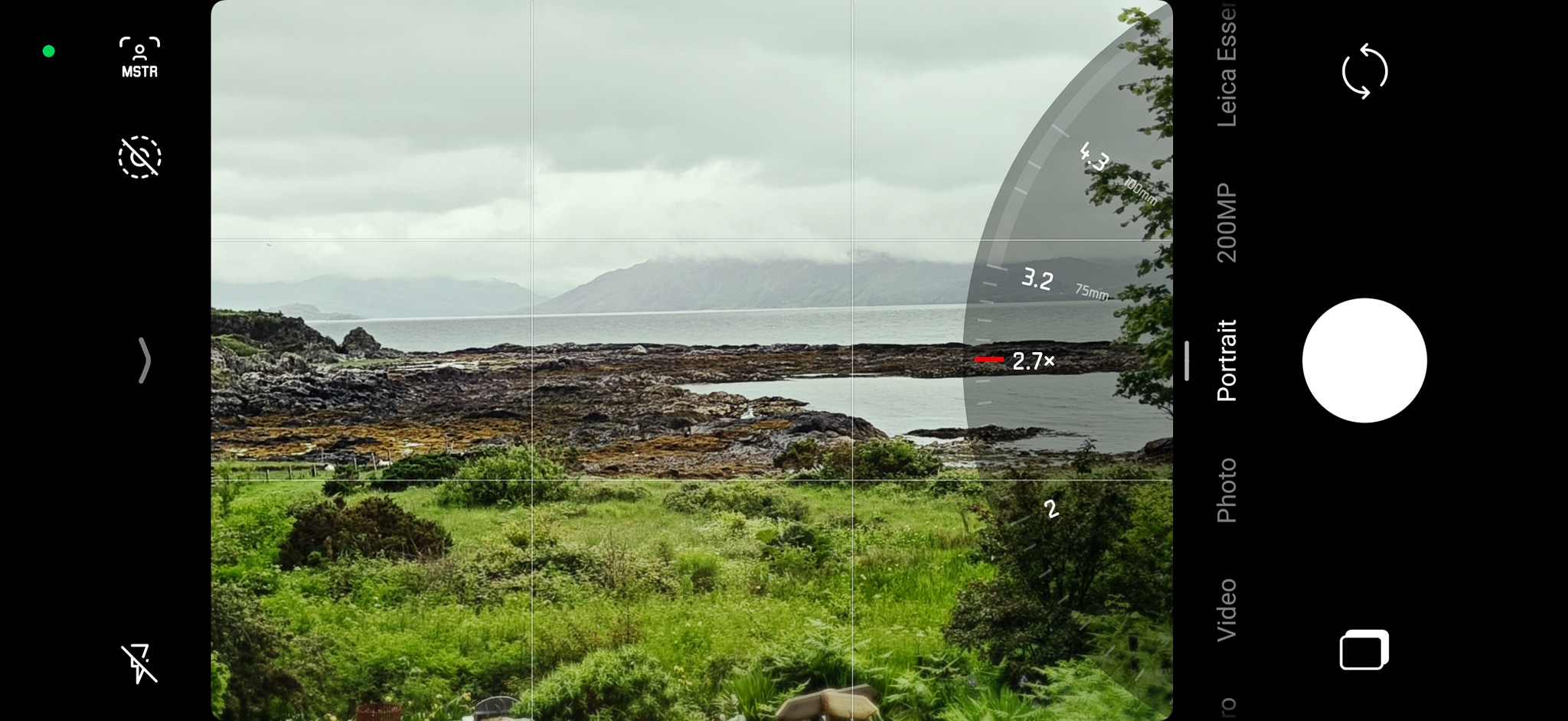 Camera viewfinder displaying a scenic coastal landscape with grey skies, rocky shores, and greenery in the foreground, featuring zoom and settings controls.