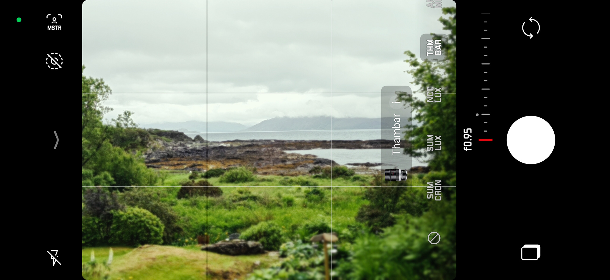 A smartphone camera interface displaying a blurred landscape of a rocky coastline and green foliage under a cloudy sky.
