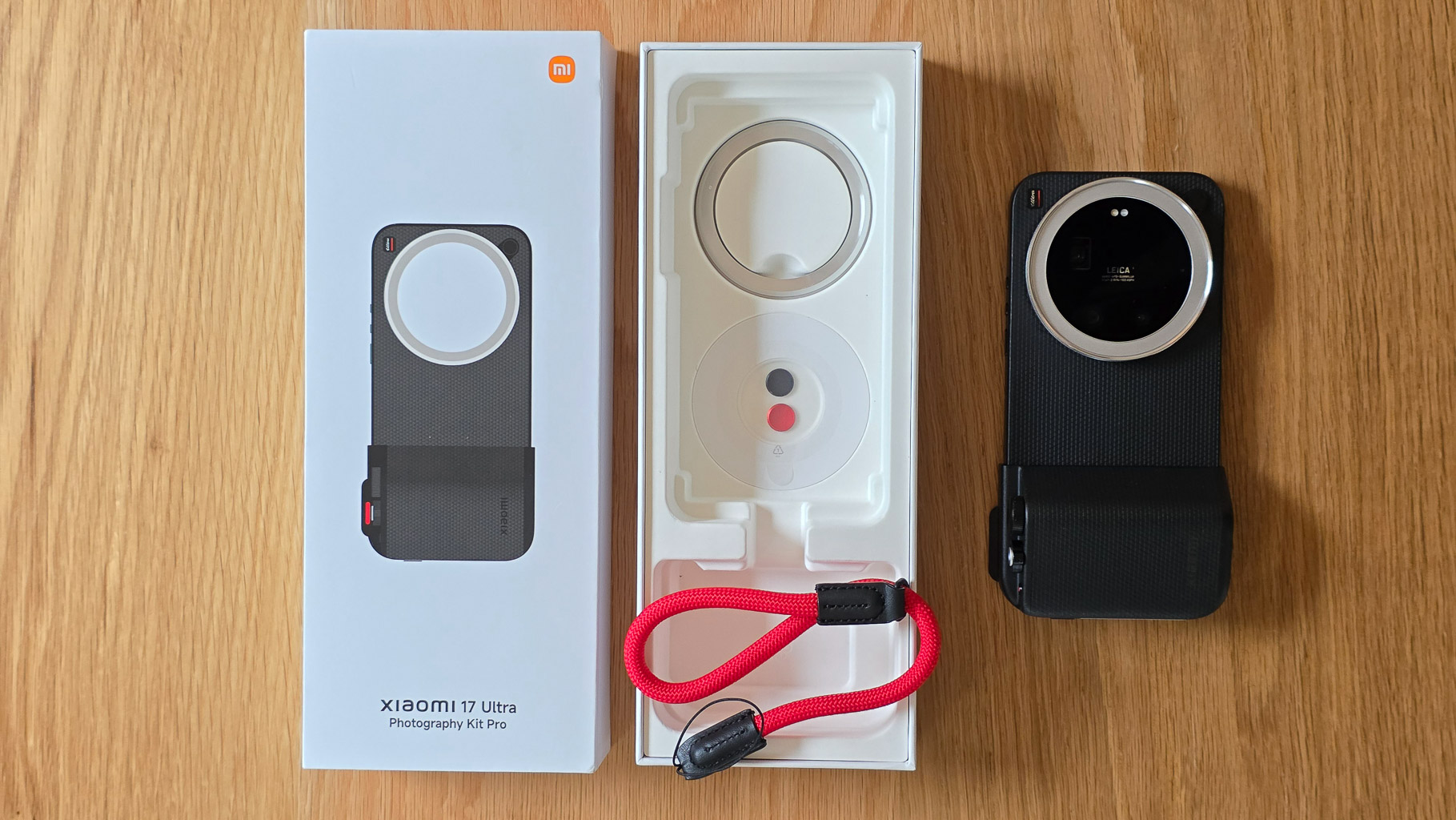 An unboxed Xiaomi 17 Ultra Photography Kit Pro displayed on a wooden surface, featuring the camera phone on the right, a white packaging box on the left, and a red camera strap in the foreground.