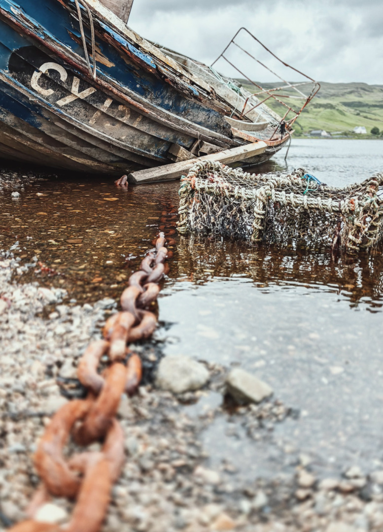 A close-up view of a rusted chain leading to an abandoned boat, partially submerged in shallow water, with a crab pot visible nearby.