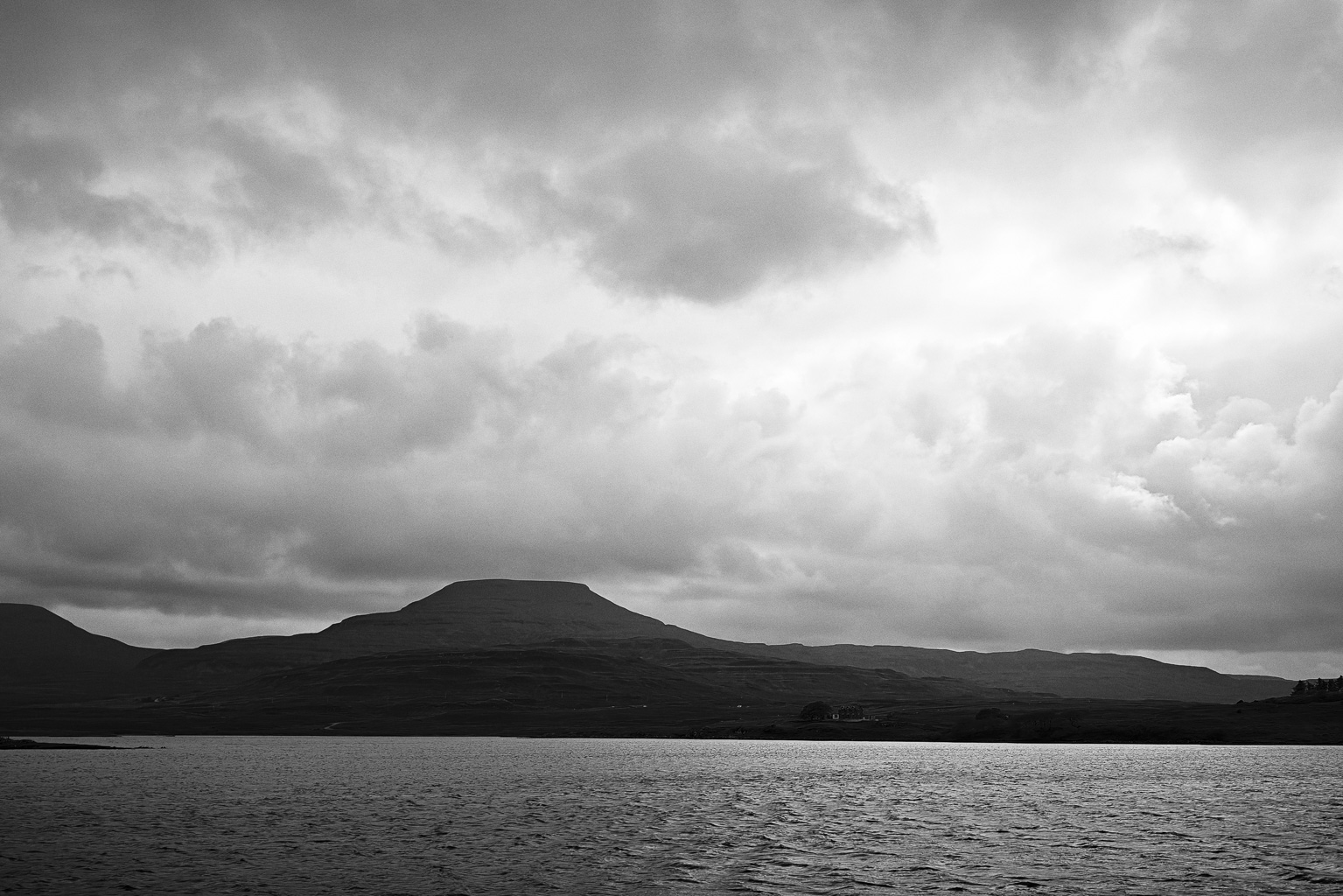 A monochrome landscape featuring a distant mountain under a cloudy sky, with a body of water in the foreground.