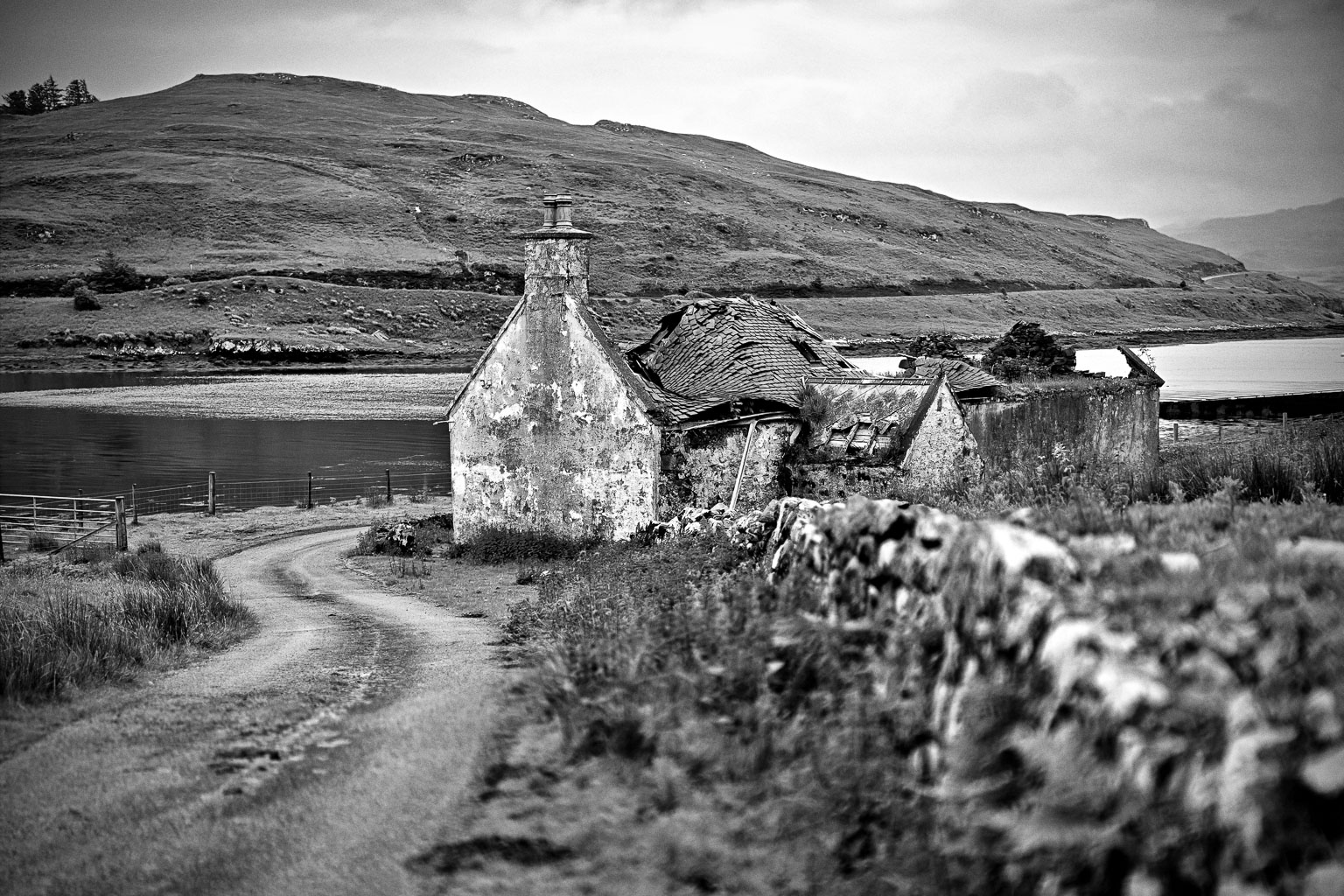 Black and white image of an old, abandoned cottage with a collapsed roof beside a winding dirt road, surrounded by grass and hills, near a body of water.