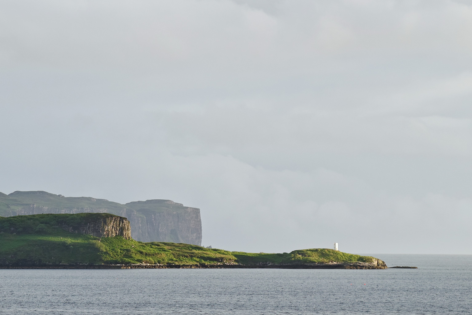 A coastal landscape featuring a green island with a lighthouse, surrounded by calm water and distant cliffs under a cloudy sky.