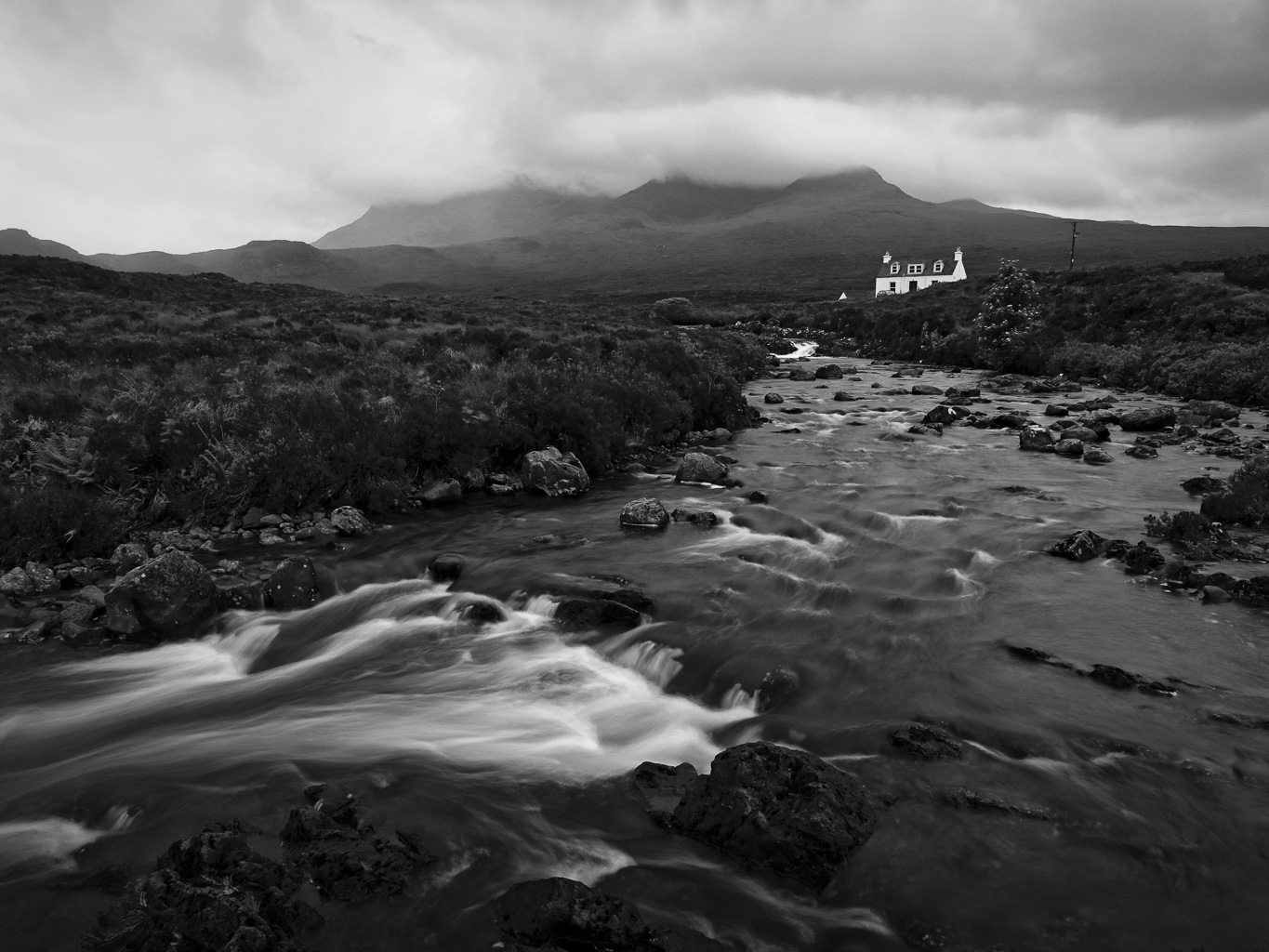 A black and white landscape featuring a flowing river, rocky banks, and a distant house set against a backdrop of mountains and overcast skies.