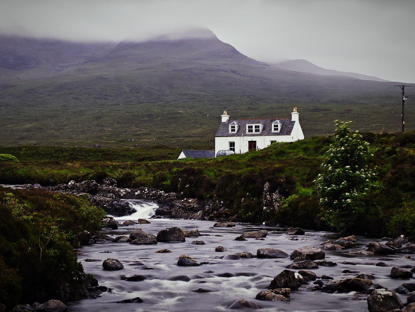 A scenic view of a white house near a flowing river, surrounded by lush greenery and mountains in the background, under a cloudy sky.