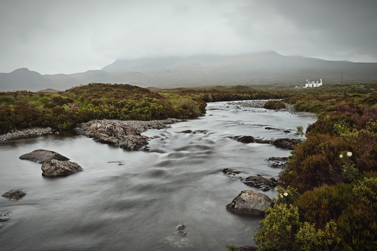 A serene landscape featuring a winding river surrounded by lush greenery and rocky terrain, with a white house in the distance and misty mountains in the background.
