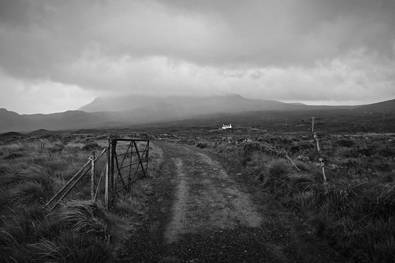 A rural dirt path leading to a distant white house, surrounded by grassy fields and mountains under a cloudy sky, depicted in black and white.