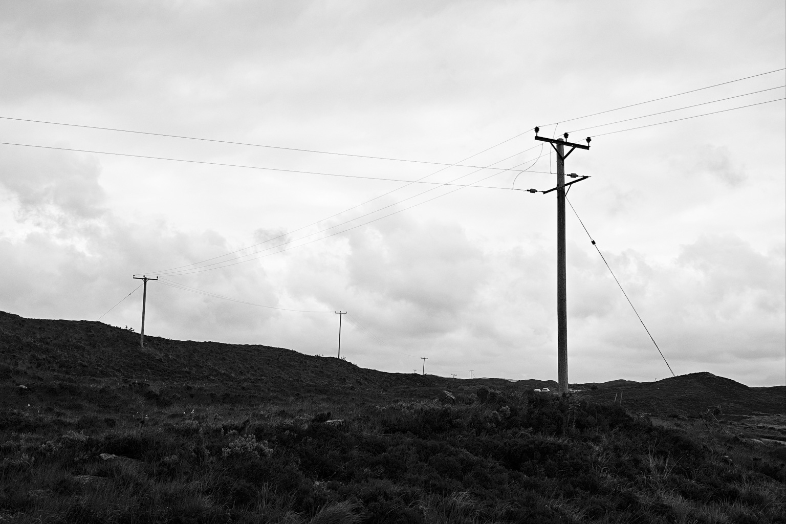 Black and white photograph of utility poles on a hilly landscape under a cloudy sky.