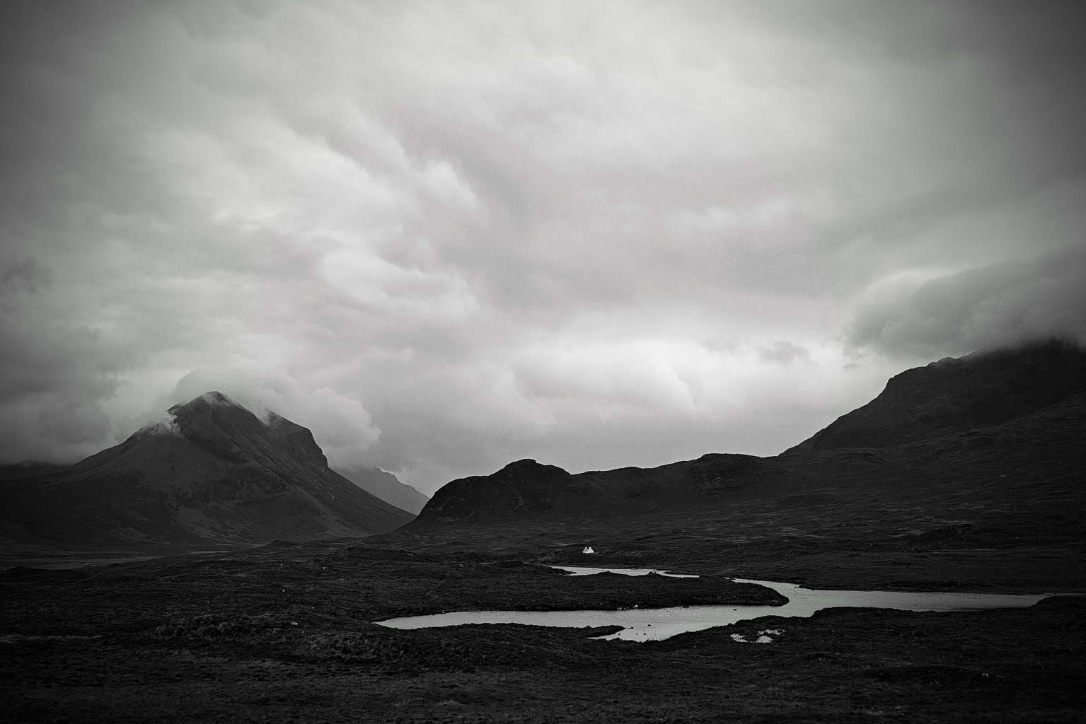 Black and white landscape featuring dramatic mountains under a cloudy sky, with a winding river and moorland in the foreground.