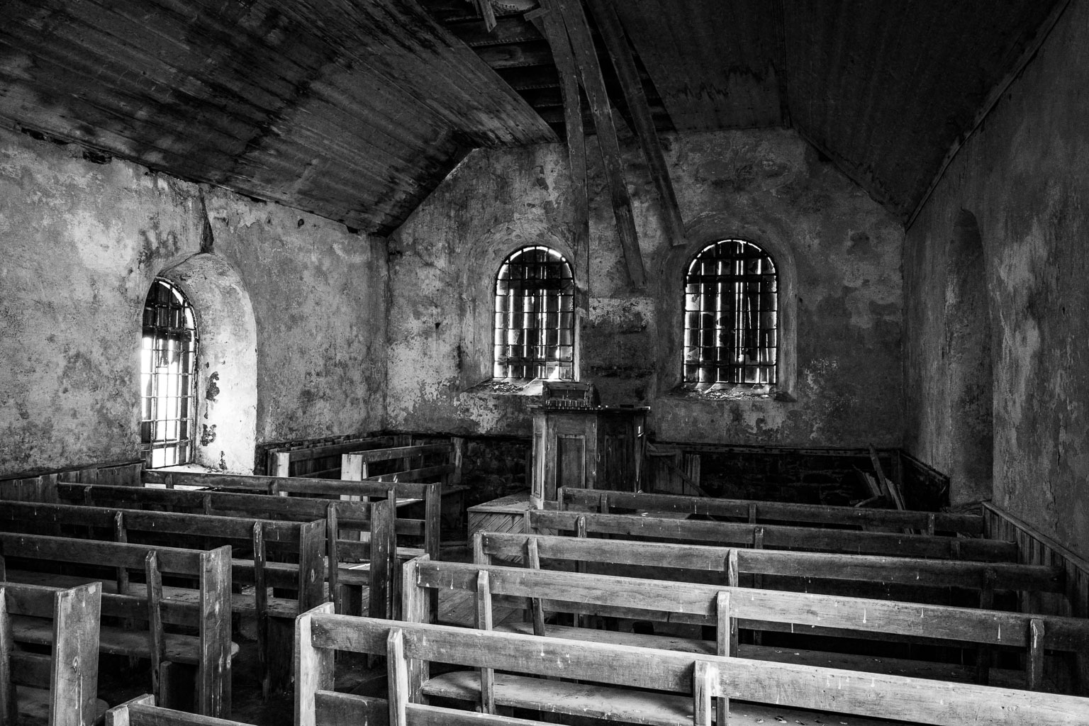 Interior of an old, abandoned church with wooden pews, cracked walls, and barred windows, captured in black and white.