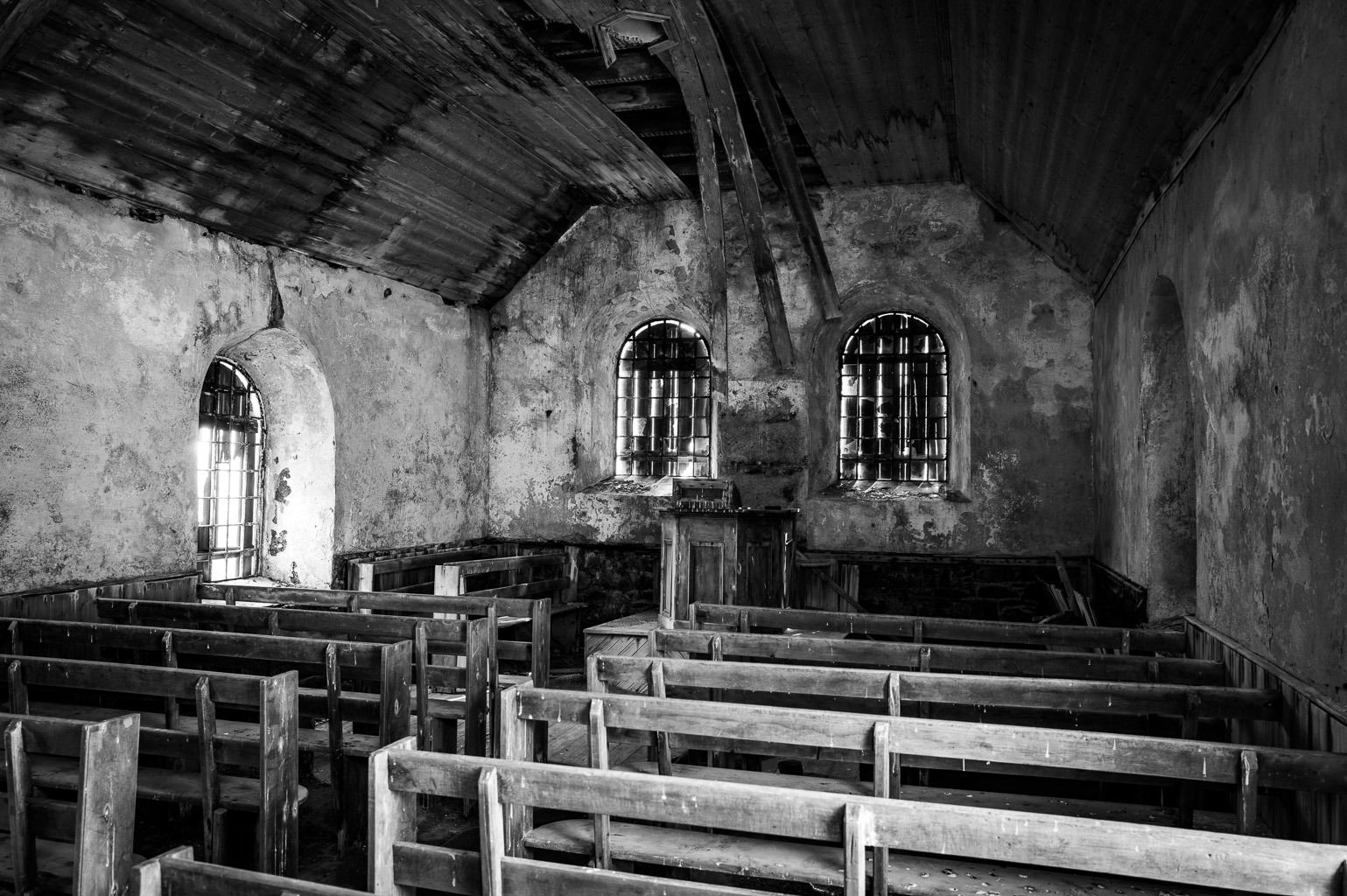 Interior of an abandoned church with wooden pews, stone walls, and arched windows, captured in black and white.