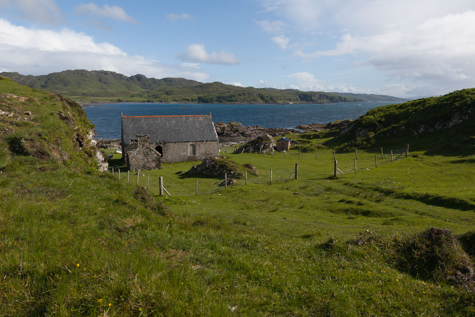 A scenic view of an old stone building near the shore, surrounded by lush green grass and hills, with a view of the ocean and distant mountains in the background.