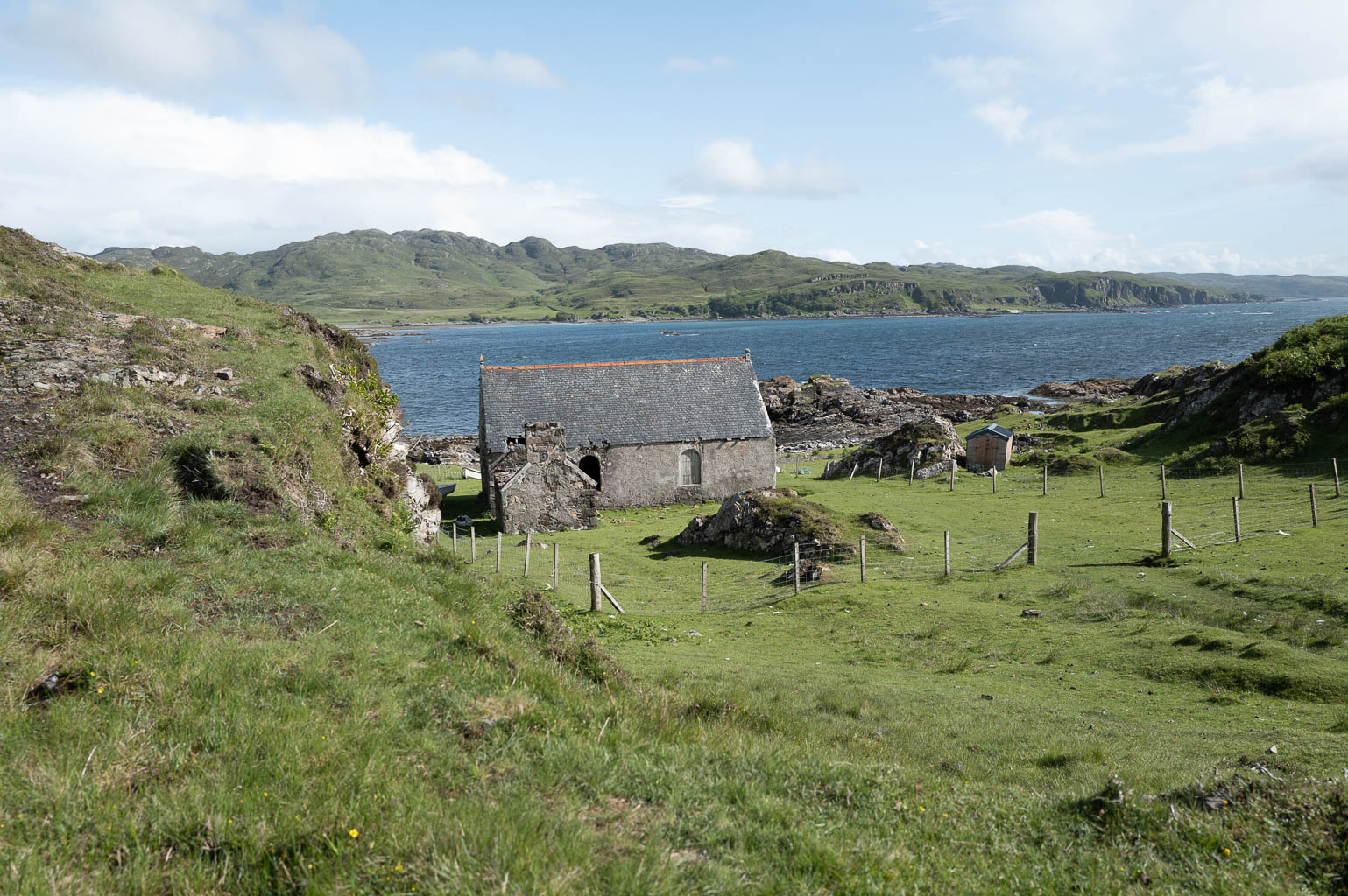 A scenic view of an old stone building near the coast, surrounded by lush green grass and hilly terrain, with a calm sea in the background.