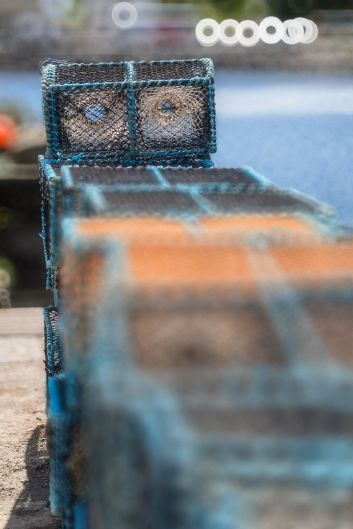 A row of lobster traps with a focus on the front trap, showcasing a blue and orange design against a blurred background of water.