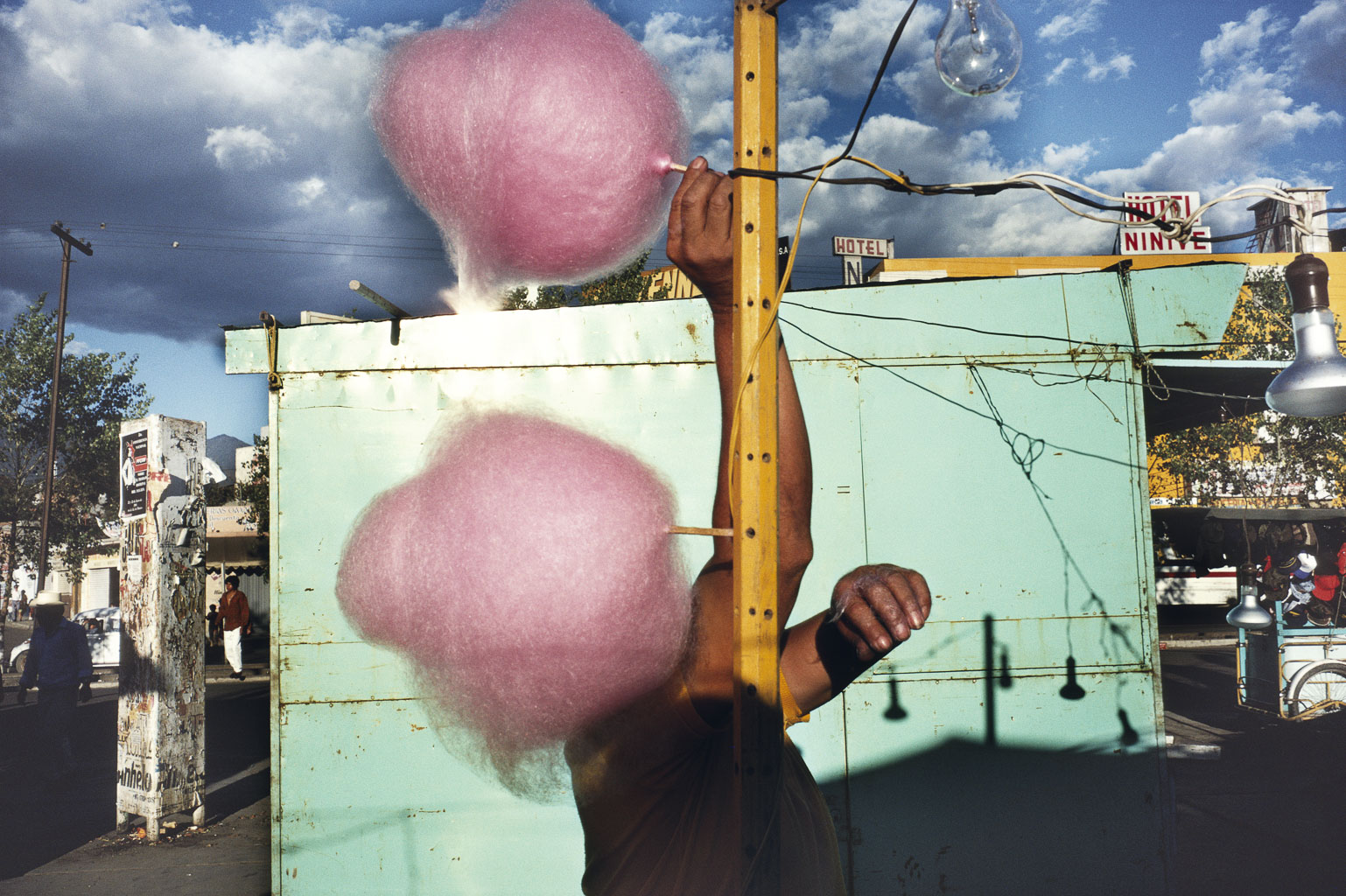 A hand holding fluffy pink cotton candy on a stick, with a vibrant outdoor setting and cloudy sky in the background. The scene includes a teal structure and visible electrical wires.