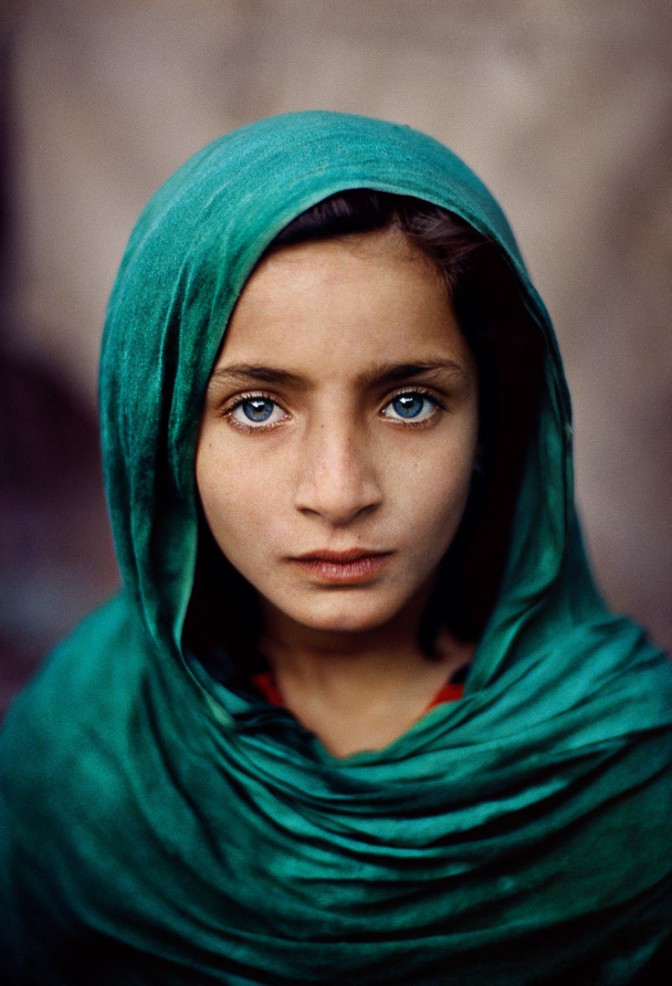 A young girl with striking blue eyes, wearing a green headscarf, gazes directly at the camera with a serious expression.
