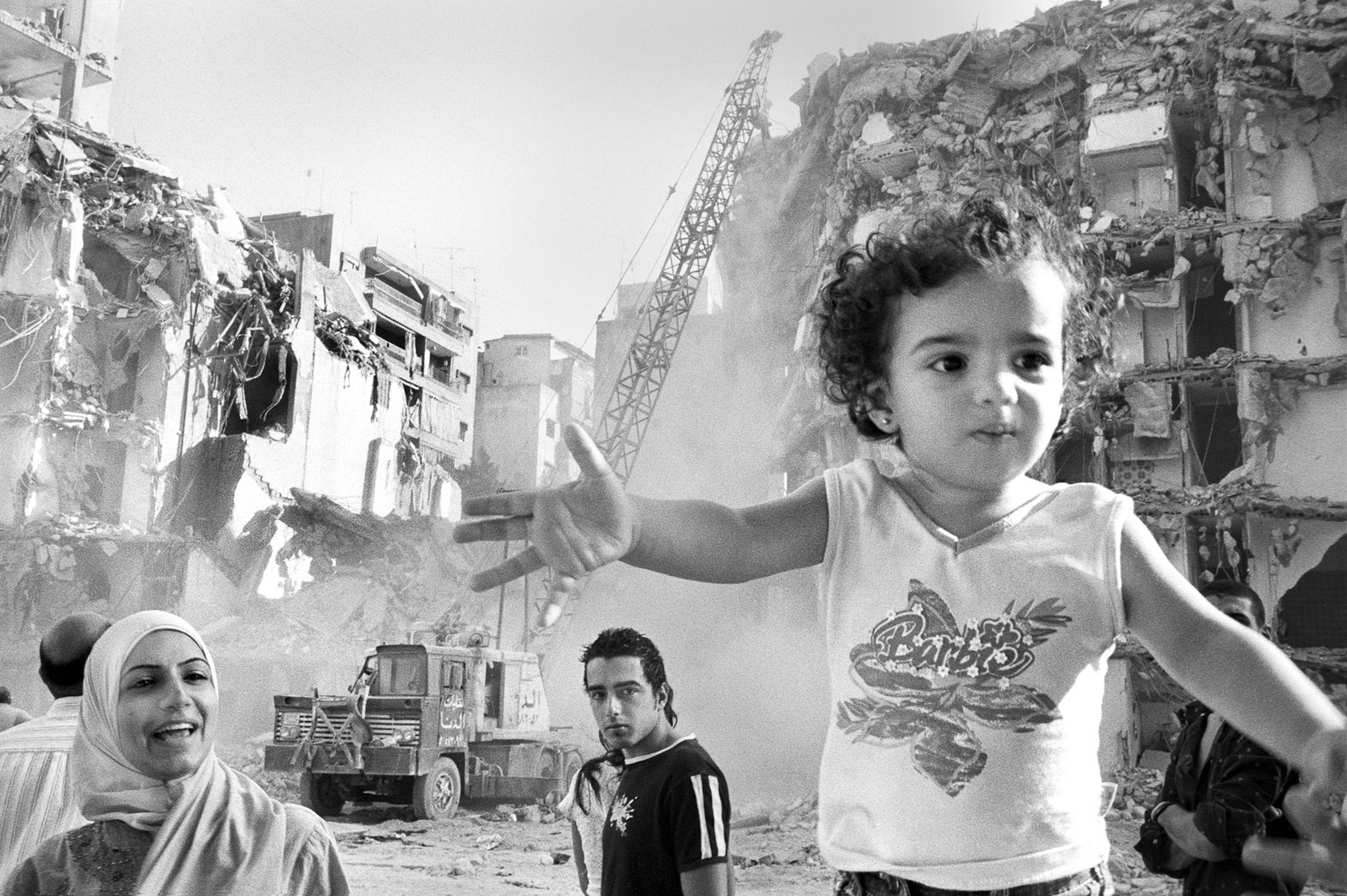 A black and white photo showing a child in a white tank top gesturing with hands, surrounded by adults, in front of a partially demolished building with construction equipment in the background.