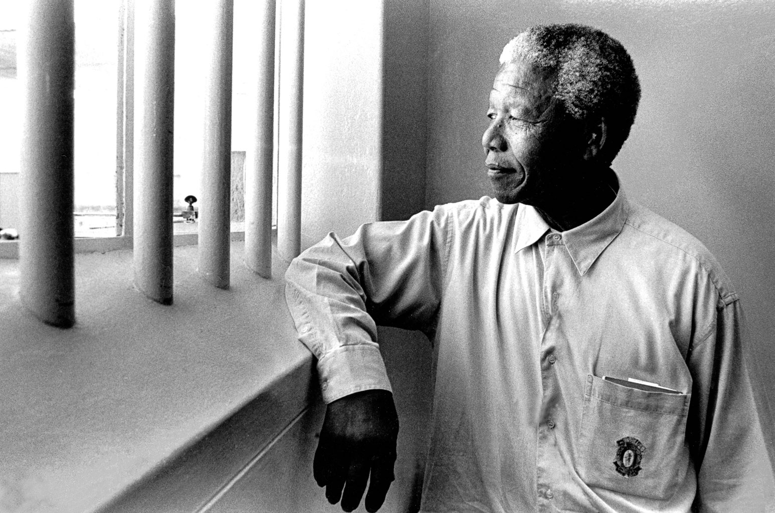 Black and white photo of a man gazing thoughtfully out of a prison window, with prison bars visible.