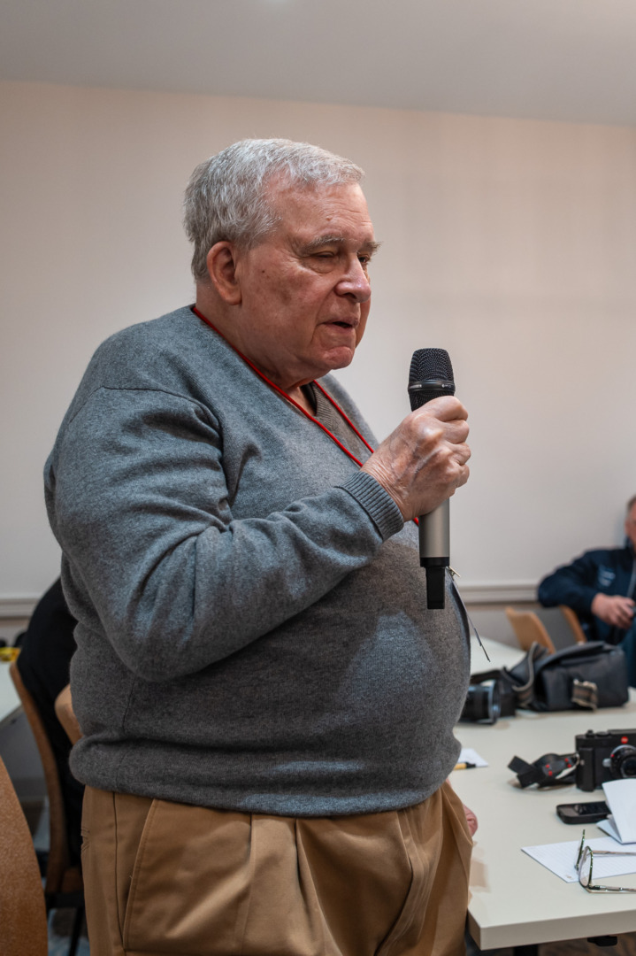 An older man with gray hair speaks into a microphone while standing in a conference room, wearing a gray sweater and tan pants.