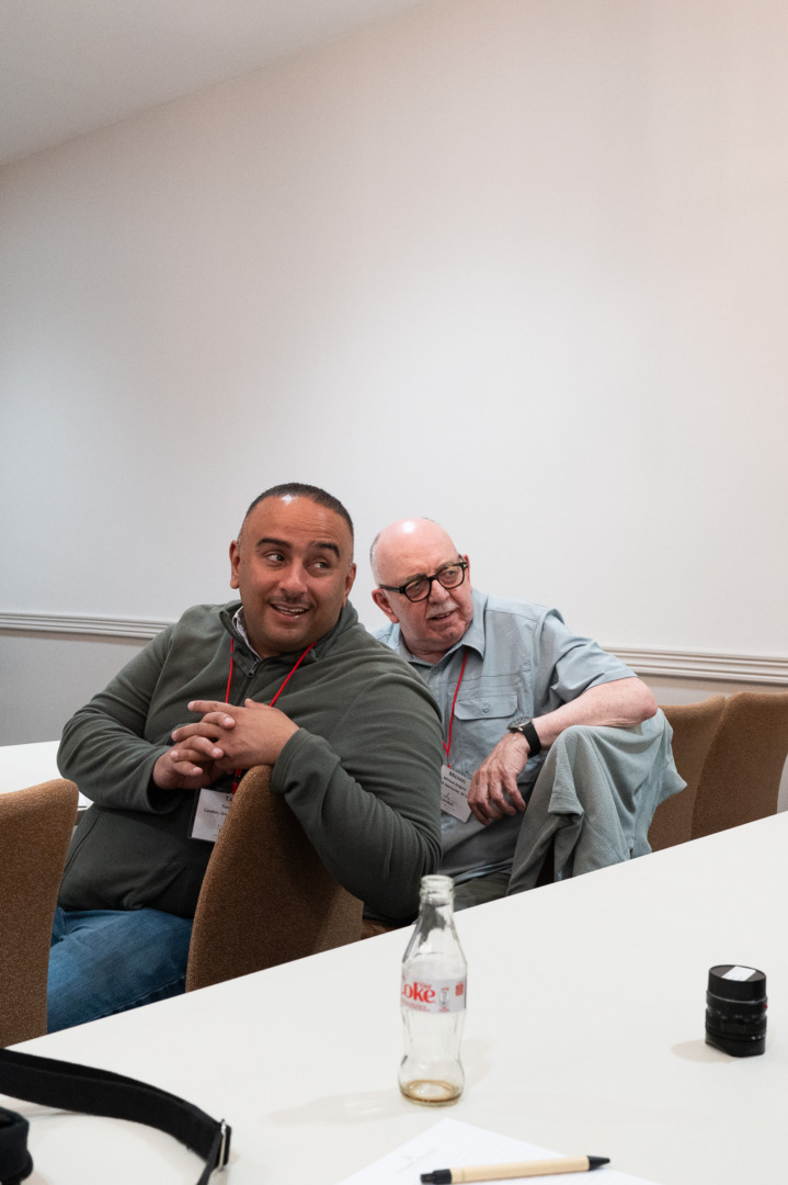 Two men sitting in a conference room, one smiling while the other looks over his shoulder. A table with a bottle and pen is visible in the foreground.