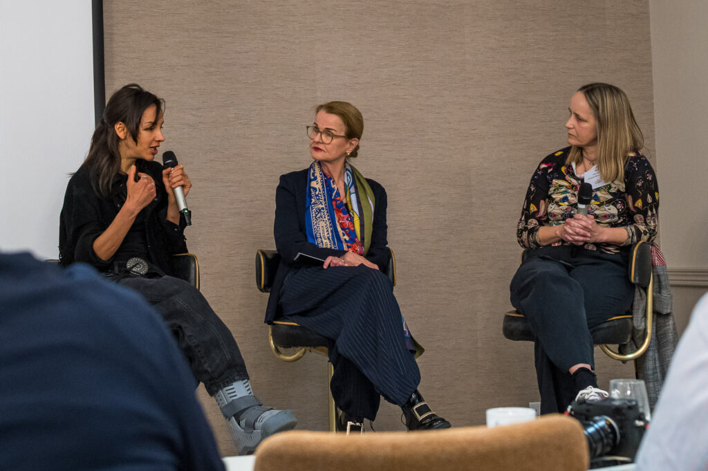 Three women participating in a panel discussion, one speaking into a microphone while the others listen attentively, seated on modern chairs.
