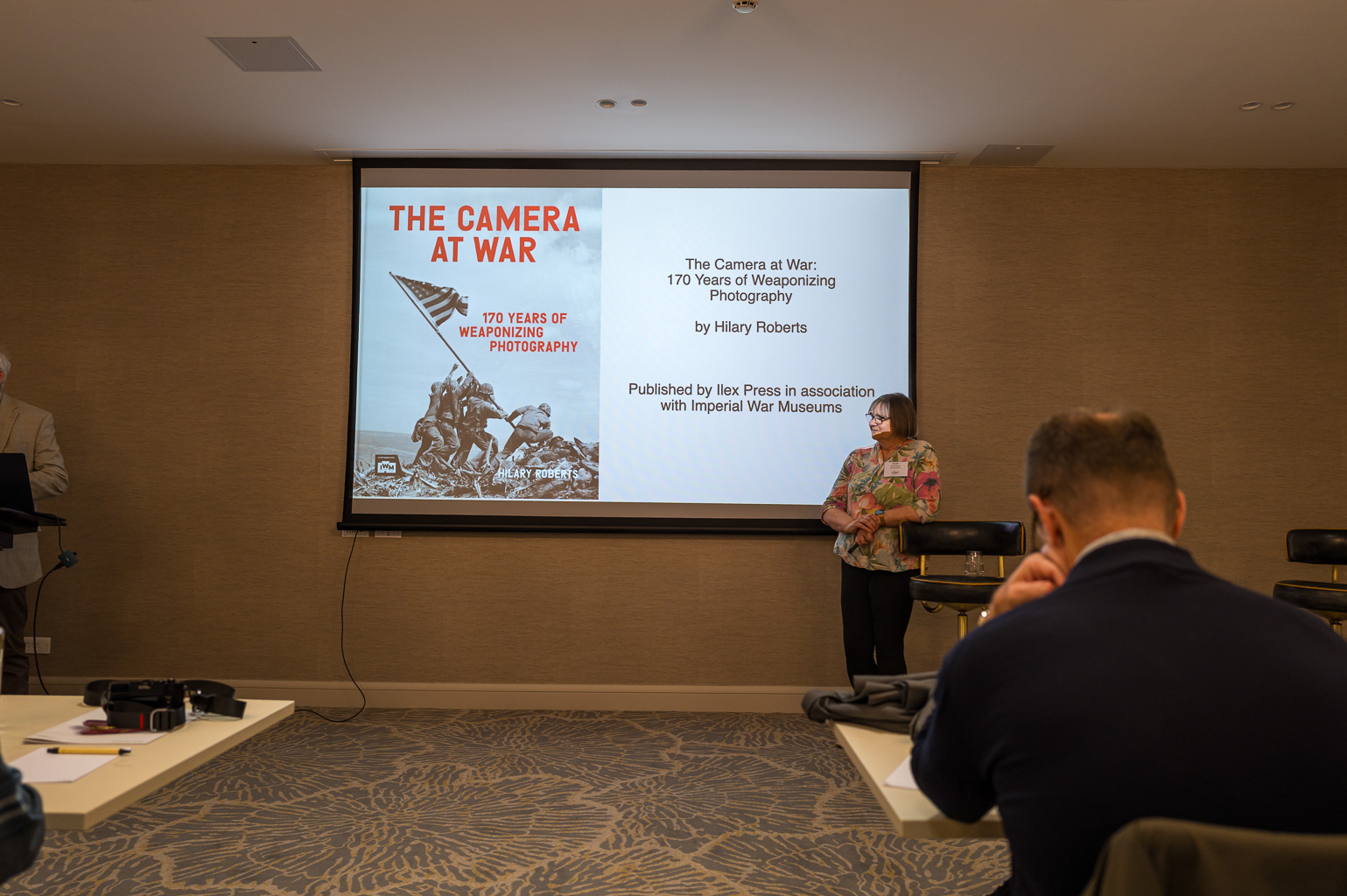 Presentation about the book 'The Camera at War: 170 Years of Weaponizing Photography' by Hilary Roberts, displayed on a screen in a conference room, with a speaker and audience members.